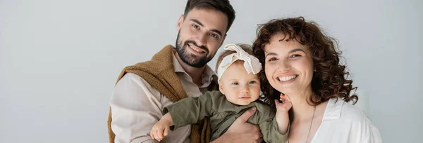 Uomo barbuto positivo che tiene in braccio la bambina in fascia vicino alla moglie riccia, striscione — Foto stock