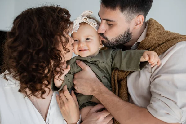 Uomo barbuto e donna riccia baciare guance di figlia felice bambino in fascia — Foto stock