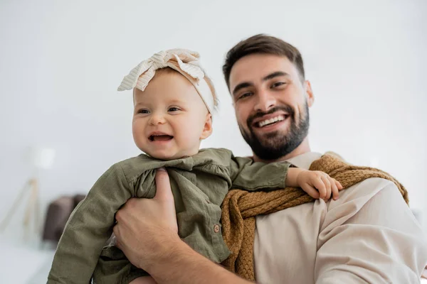 Cheerful bearded man holding in arms infant girl in headband and looking at camera — Stock Photo