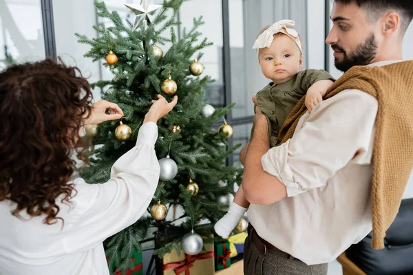Curly woman decorating christmas tree near bearded husband and infant daughter — Stock Photo