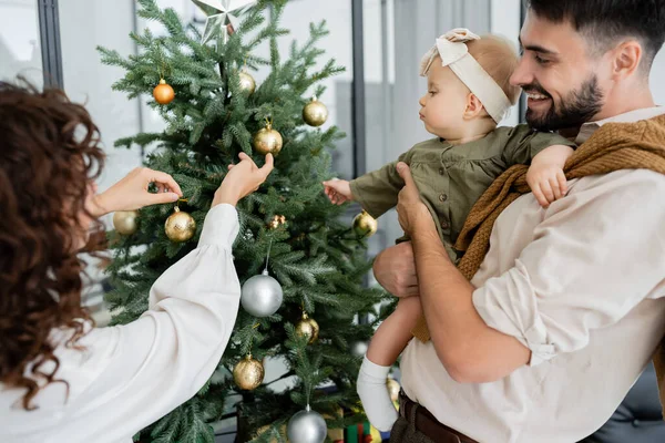Lockige Frau schmückt Weihnachtsbaum in der Nähe von glücklichem Ehemann und kleiner Tochter — Stockfoto