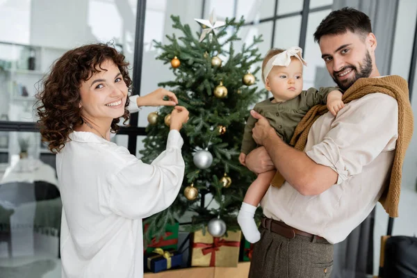 Mujer alegre decorando árbol de Navidad cerca de marido feliz e hija bebé - foto de stock