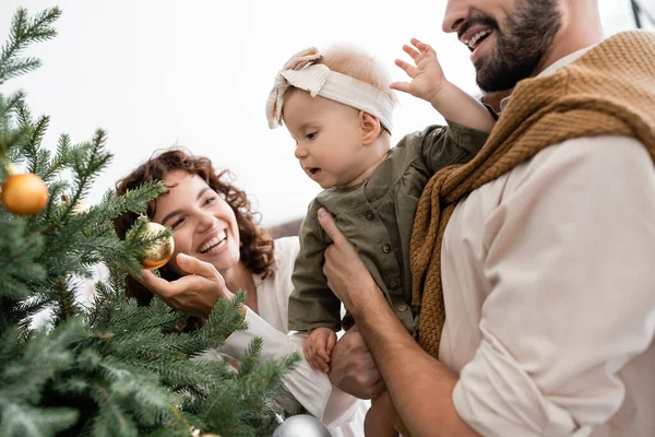 Bärtiger Mann hält kleine Tochter in der Nähe von geschmücktem Weihnachtsbaum und fröhlicher Frau — Stockfoto