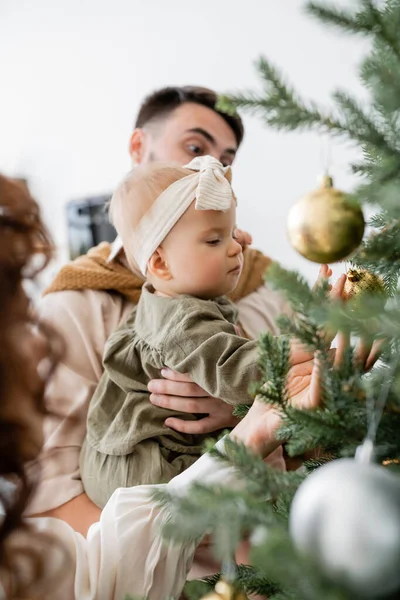 Father holding infant daughter near decorated christmas tree and curly wife — Stock Photo