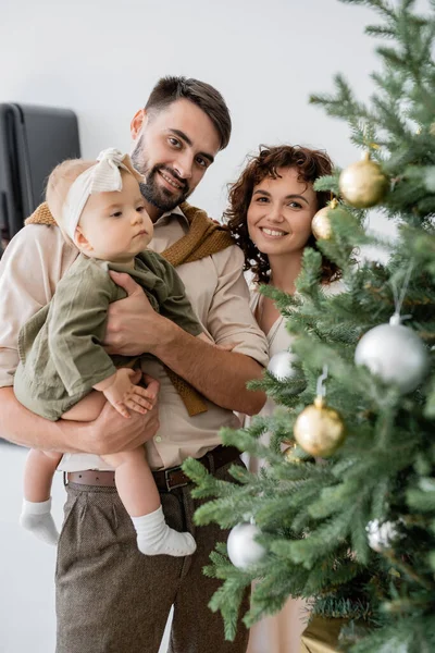 Glücklicher Vater hält kleine Tochter neben fröhlicher Frau und geschmücktem Weihnachtsbaum — Stockfoto