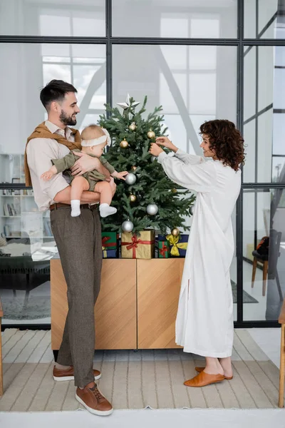 Curly woman decorating christmas tree near bearded husband and baby daughter — Stock Photo