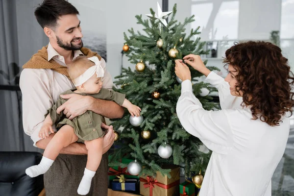 Mujer feliz decorando árbol de Navidad cerca de marido barbudo e hija bebé - foto de stock