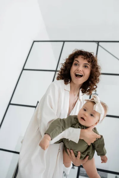 Excited and curly woman in white dress holding infant daughter in arms — Stock Photo