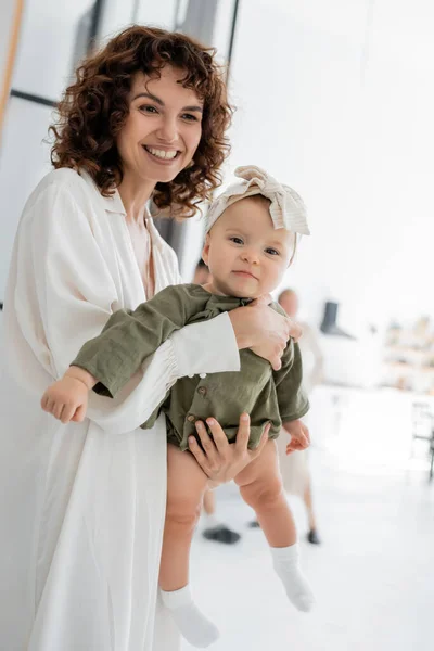 Cheerful and curly woman in white dress holding infant daughter in arms — Stock Photo