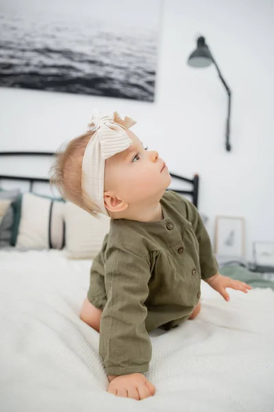 Curious baby girl in dress and headband with bow sitting on bed at home — Stock Photo