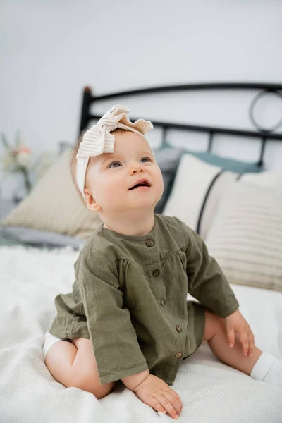 Cheerful baby girl in dress and headband with bow sitting on bed at home — Stock Photo