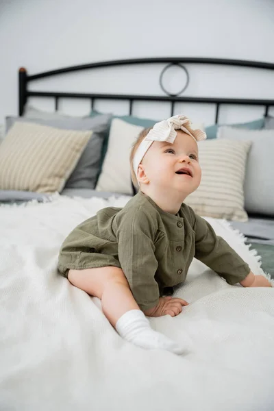 Happy baby girl in dress and headband with bow sitting on bed at home — Stock Photo