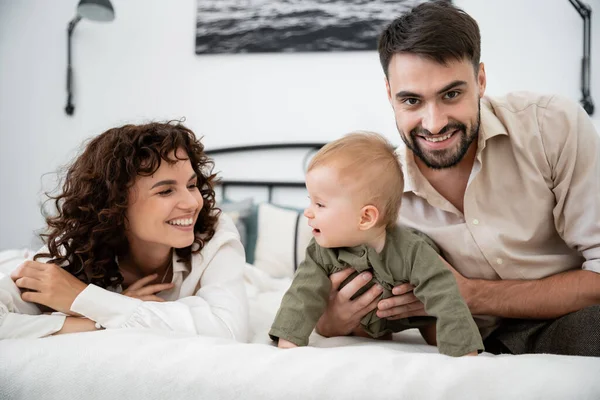 Glückliche Mutter sieht Baby-Mädchen neben fröhlichem Ehemann auf dem Bett an — Stockfoto