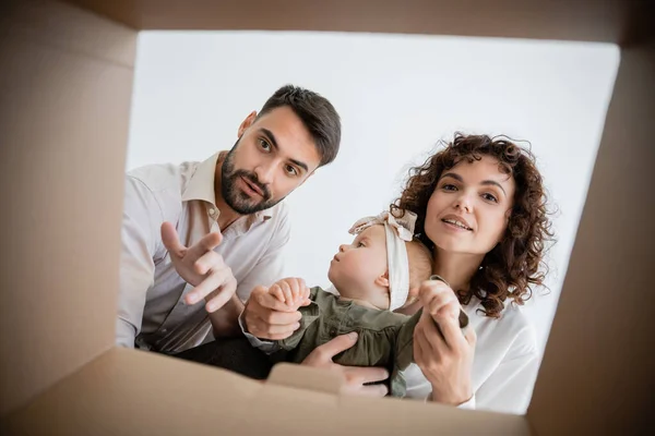 Vista inferior de los padres curiosos sosteniendo bebé niña y mirando dentro de la caja de cartón - foto de stock