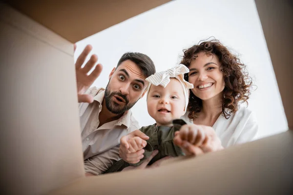 Vista inferior de padres alegres y niña linda bebé mirando dentro de la caja de cartón - foto de stock