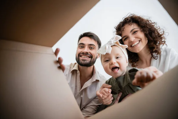 Vista inferior de padres alegres y niña bebé asombrada mirando dentro de la caja de cartón - foto de stock