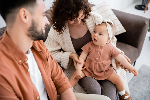 Lockige Mutter hält ihre kleine Tochter in der Nähe eines bärtigen Mannes, der auf der Couch sitzt — Stockfoto