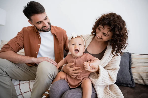 Cheerful mother holding amazed infant daughter near bearded husband sitting on couch — Stock Photo