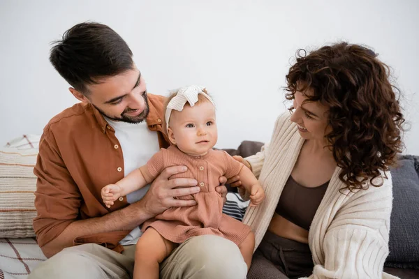 Cheerful father holding happy infant daughter near curly wife sitting on couch — Stock Photo