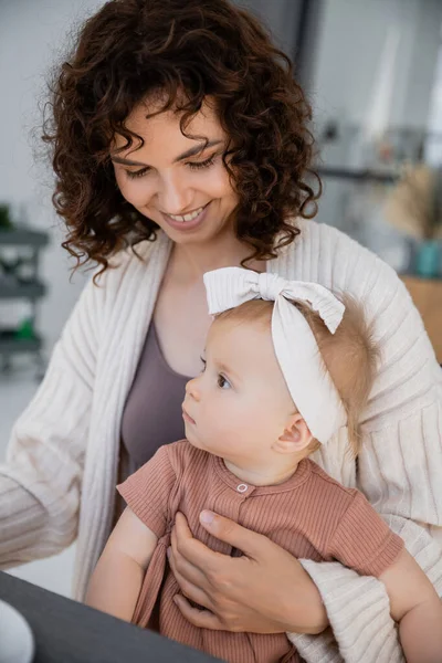 Madre positiva con i capelli ricci sorridenti mentre tiene la bambina in fascia — Foto stock