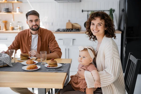 Allegra madre che tiene la figlia neonata e si siede a tavola con il marito durante la colazione — Foto stock