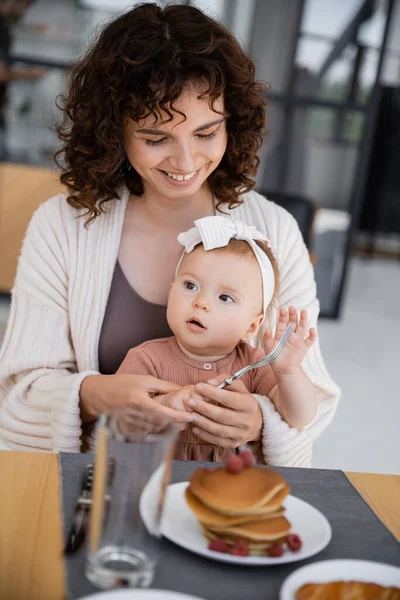 Mulher feliz sentado com bebê filha segurando garfo perto de panquecas com framboesas — Fotografia de Stock