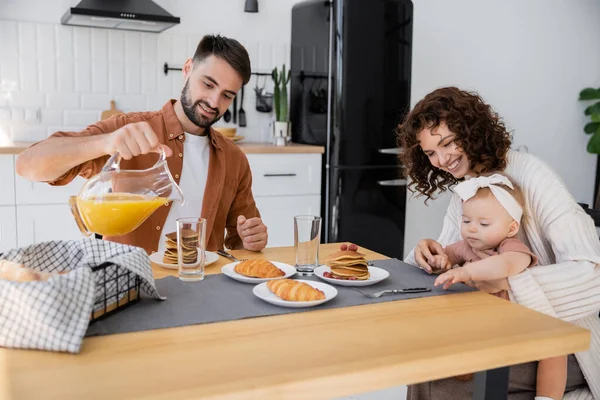 Бородатый мужчина наливает апельсиновый сок возле счастливой семьи во время завтрака — стоковое фото