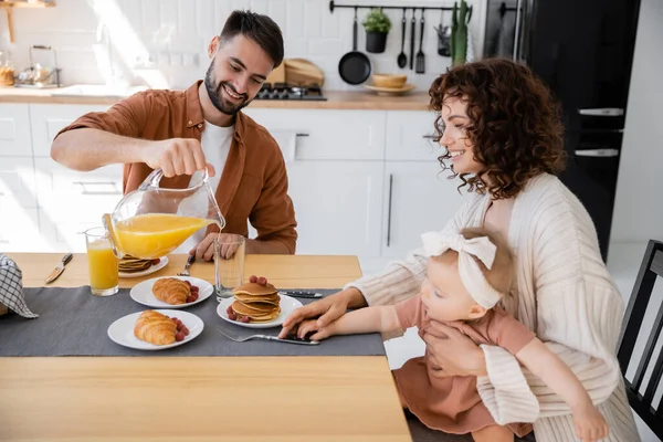 Bärtiger Mann gießt beim Frühstück Orangensaft in die Nähe von glücklicher Frau und kleiner Tochter — Stockfoto