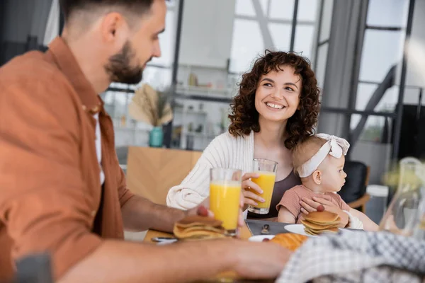 Счастливая женщина, держащая младенческую дочь и смотрящая на босого мужа во время завтрака — стоковое фото