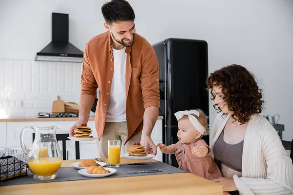 Bärtiger Mann serviert seiner glücklichen Frau mit kleiner Tochter beim Frühstück Pfannkuchen — Stockfoto