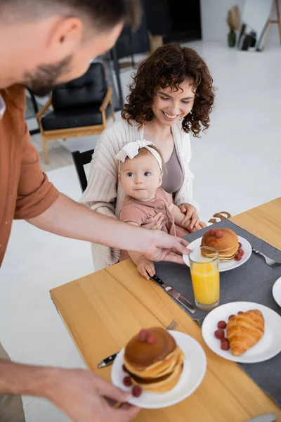 Мужчина подает блинчики счастливой жене с младенческой дочерью во время завтрака — стоковое фото