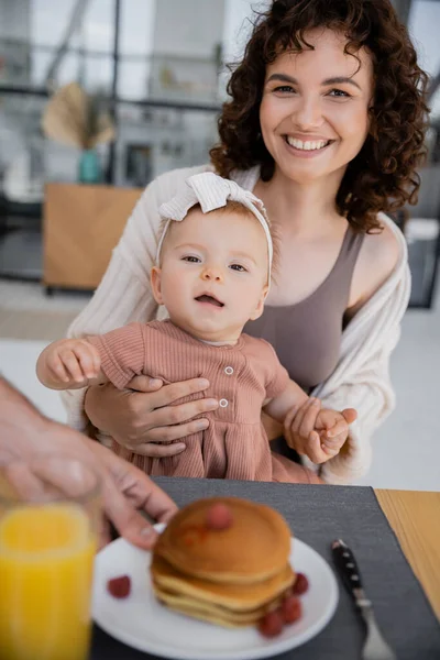 Uomo che serve frittelle alla moglie allegra con figlia neonata durante la colazione — Foto stock