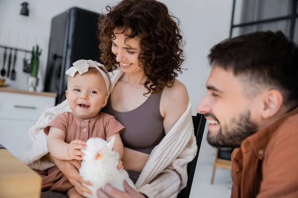 Happy bearded man holding soft toy near happy baby daughter and curly wife — Stock Photo