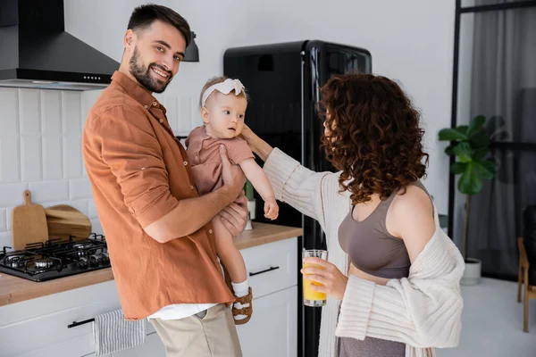 Alegre hombre sosteniendo bebé hija cerca rizado esposa con vaso de jugo de naranja fresca en cocina - foto de stock