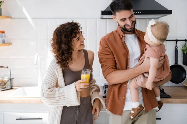 Uomo soddisfatto che tiene la figlia del bambino vicino alla moglie riccia con un bicchiere di succo d'arancia fresco in cucina — Foto stock