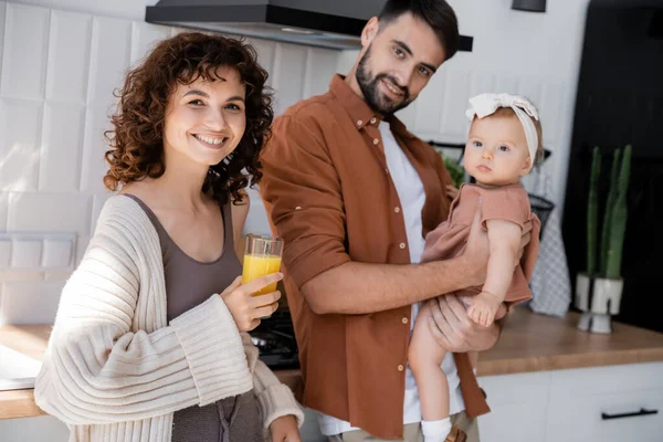 Alegre hombre sosteniendo bebé hija cerca rizado esposa con vaso de jugo de naranja fresco en cocina - foto de stock