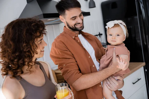 Happy man holding baby daughter near curly wife with glass of fresh orange juice in kitchen — Stock Photo