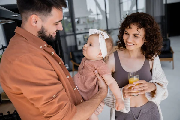 Pai feliz segurando bebê filha perto da esposa encaracolada com vidro de suco de laranja fresco na cozinha — Fotografia de Stock