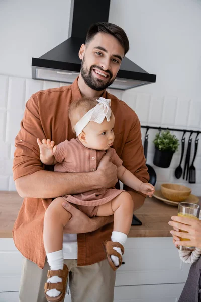 Feliz padre sosteniendo bebé hija cerca de esposa con vaso de jugo de naranja fresco en la cocina - foto de stock