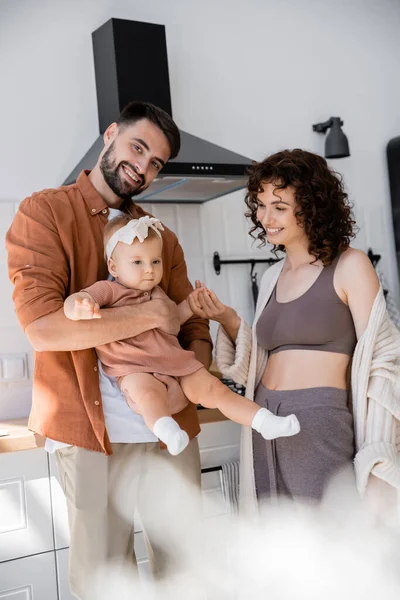 Homem barbudo feliz segurando nos braços criança filha perto sorrindo esposa na cozinha — Fotografia de Stock