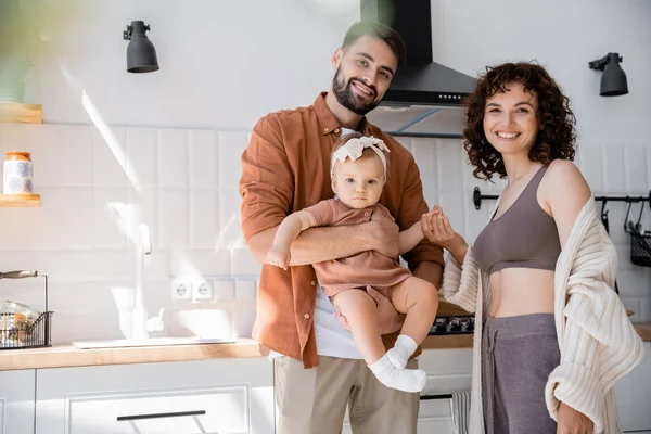 Heureux barbu homme tenant dans les bras bébé fille près joyeuse femme dans la cuisine — Photo de stock