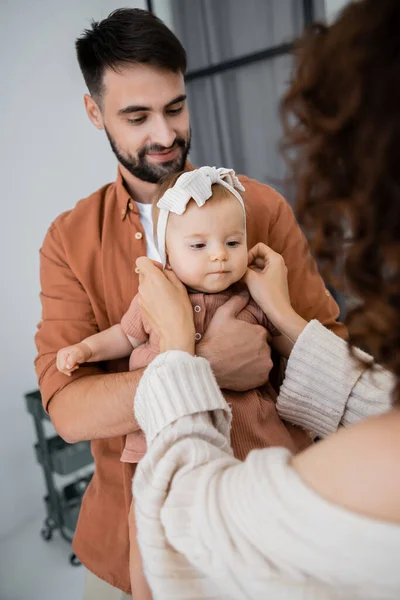 Curly mother adjusting headband of baby daughter near cheerful husband at home — Stock Photo