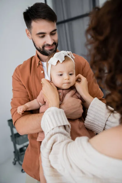 Mãe encaracolado ajustando a cabeça da filha do bebê perto do marido alegre em casa — Fotografia de Stock