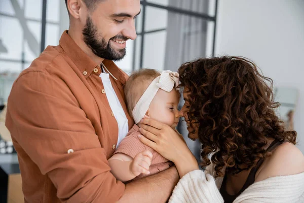 Curly mother touching nose of baby daughter in headband near cheerful husband at home — Stock Photo