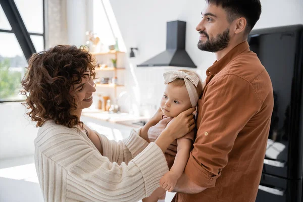 Felice padre barbuto che tiene in braccio la figlia del bambino vicino alla moglie compiaciuta a casa — Foto stock