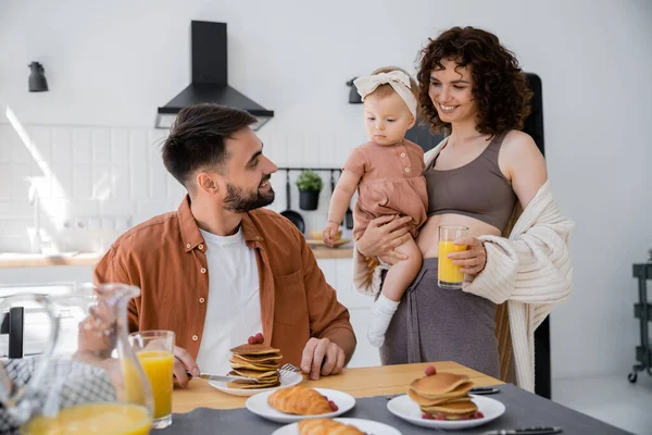 Cheerful woman holding in arms baby daughter and bringing orange juice to husband during breakfast — Stock Photo