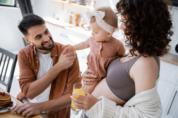 Curly woman holding in arms baby daughter and bringing orange juice to husband during breakfast — Stock Photo