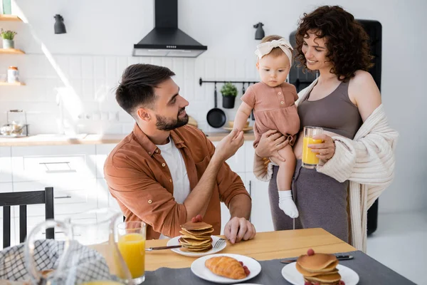 Femme gaie tenant dans les bras fille enfant et apportant du jus d'orange au mari pendant le petit déjeuner — Photo de stock