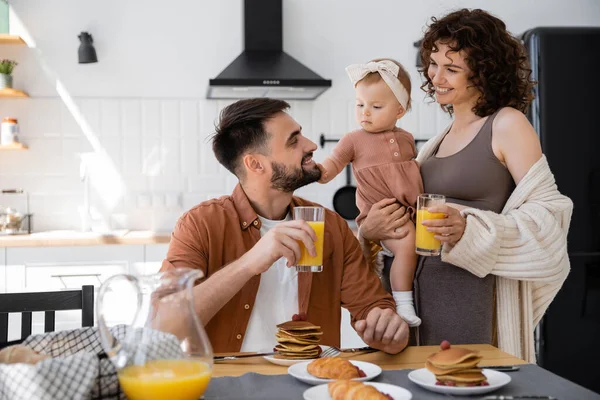 Happy parents holding glasses of orange juice near infant baby during breakfast — Stock Photo