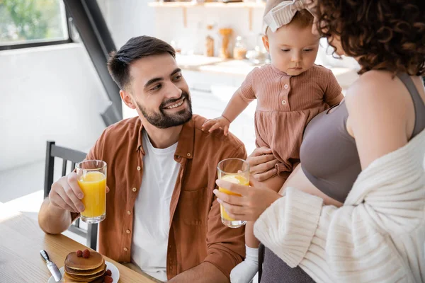 Mulher encaracolado segurando nos braços bebê infantil perto marido sorridente com copo de suco de laranja — Fotografia de Stock